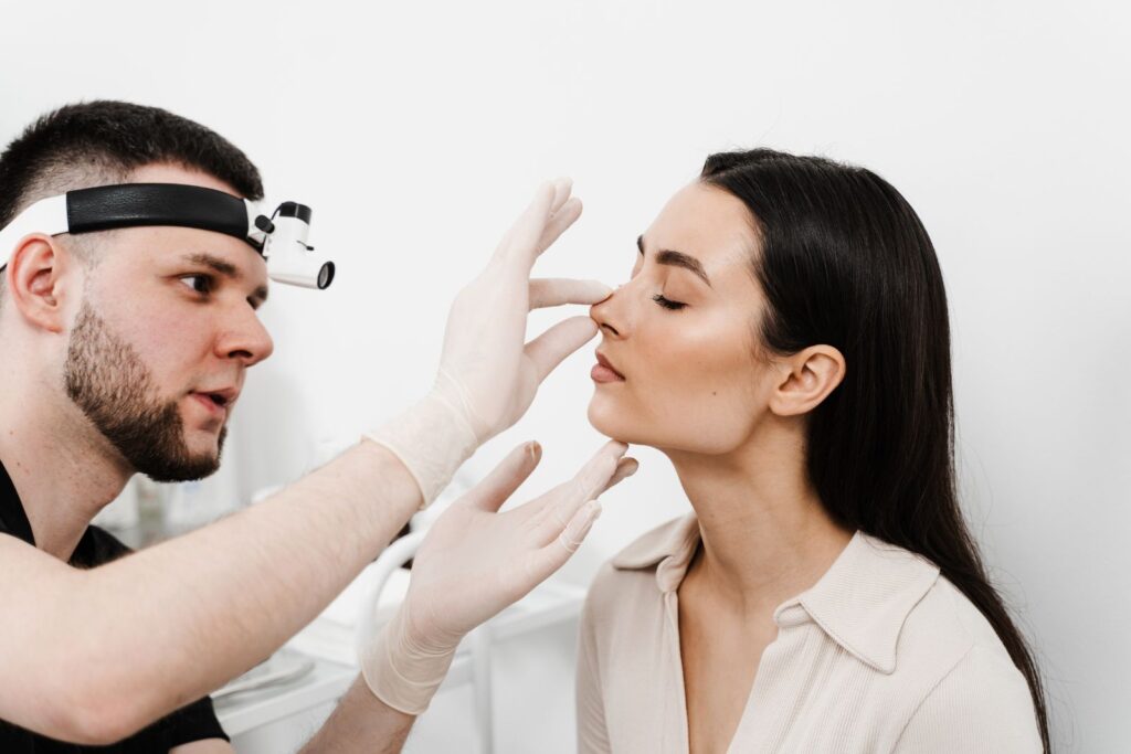 A medical professional examines a patient's nose in a clinical setting. The patient's eyes are closed, and the doctor wears gloves and a head magnifier.