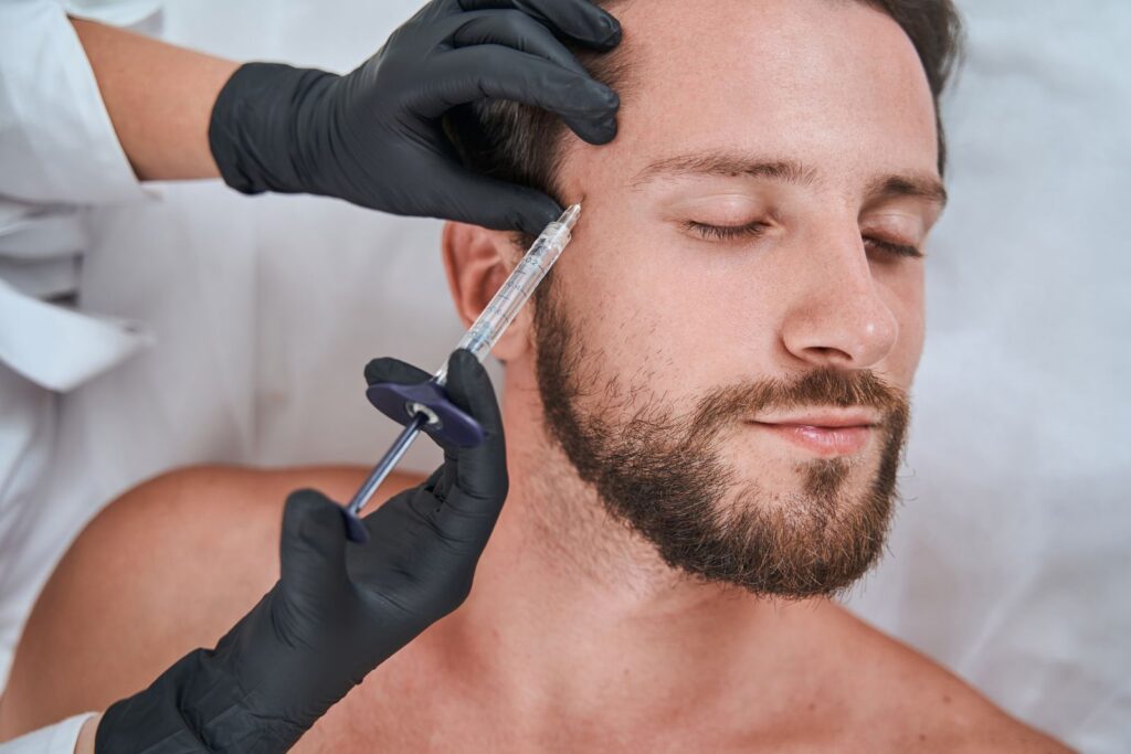 A person with a beard receives a facial injection from a professional wearing black gloves, skillfully administering one of the many types of dermal fillers.