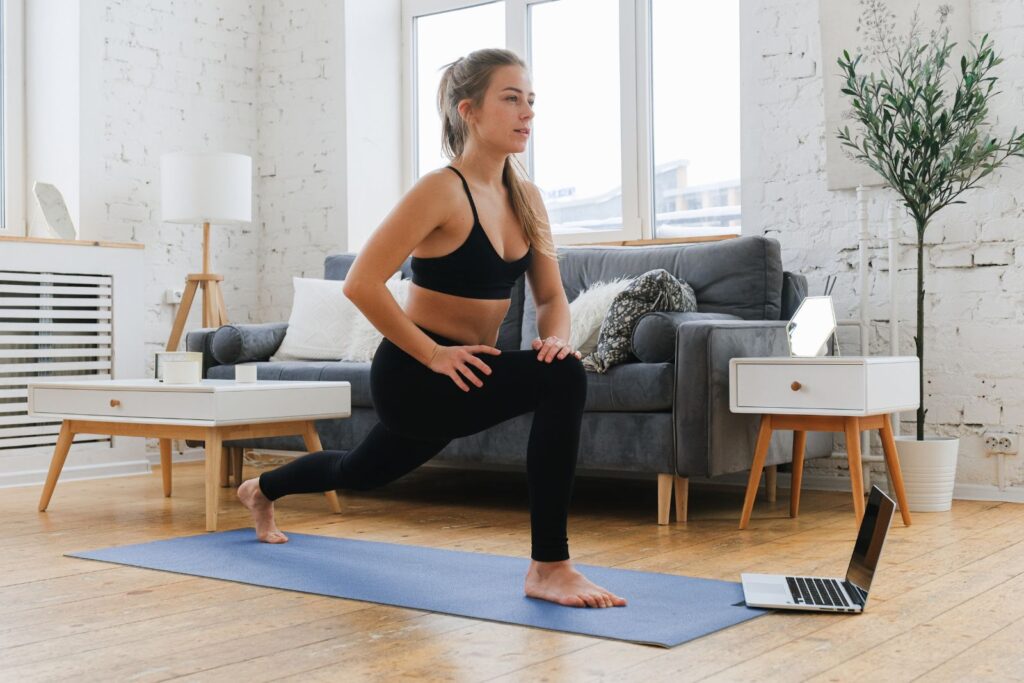A woman in workout attire does lunges on a yoga mat in her living room, facing an open laptop on the floor. She's researching what BPC 157 is to enhance her fitness routine.