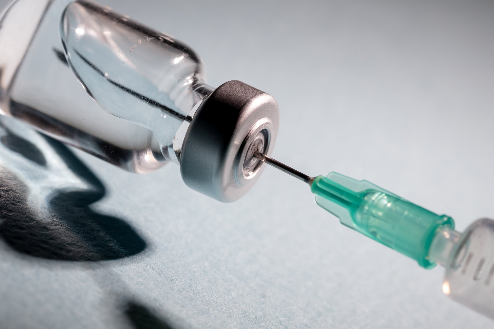 A syringe with a green tip needle pierces the rubber stopper of a clear vial containing BPC 157, set against a light blue background.