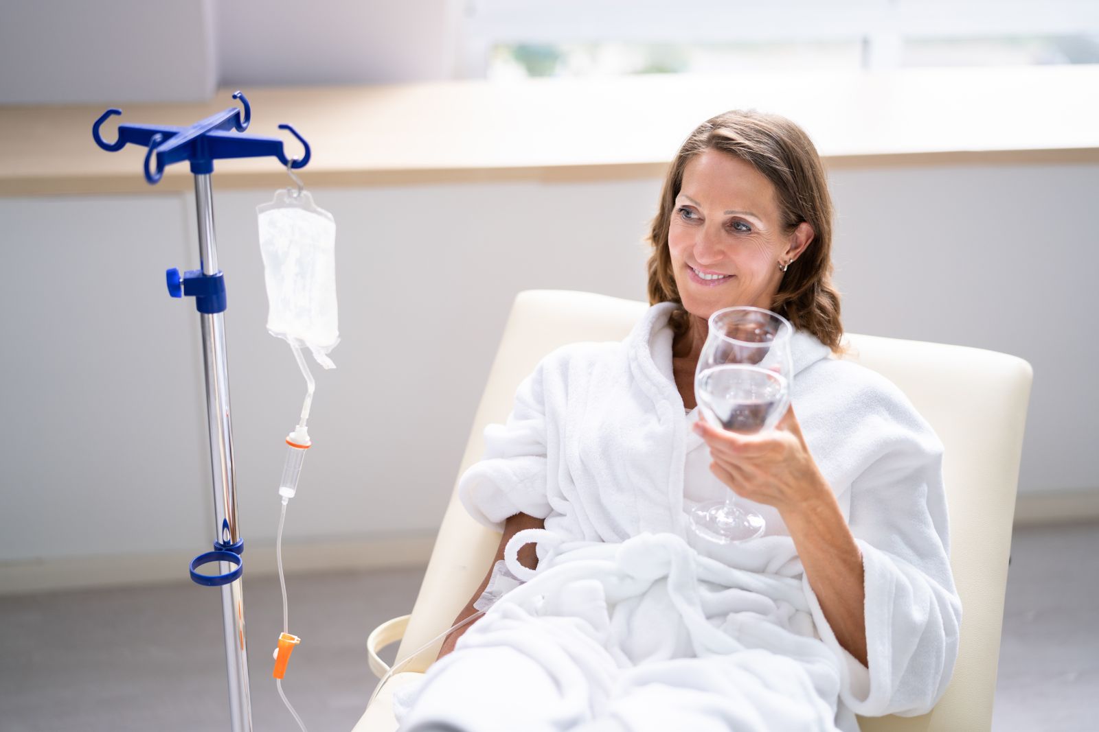 A woman in a white robe sits in a medical chair, holding a glass of water next to an IV drip stand in a bright room, receiving a NAD anti-aging treatment.
