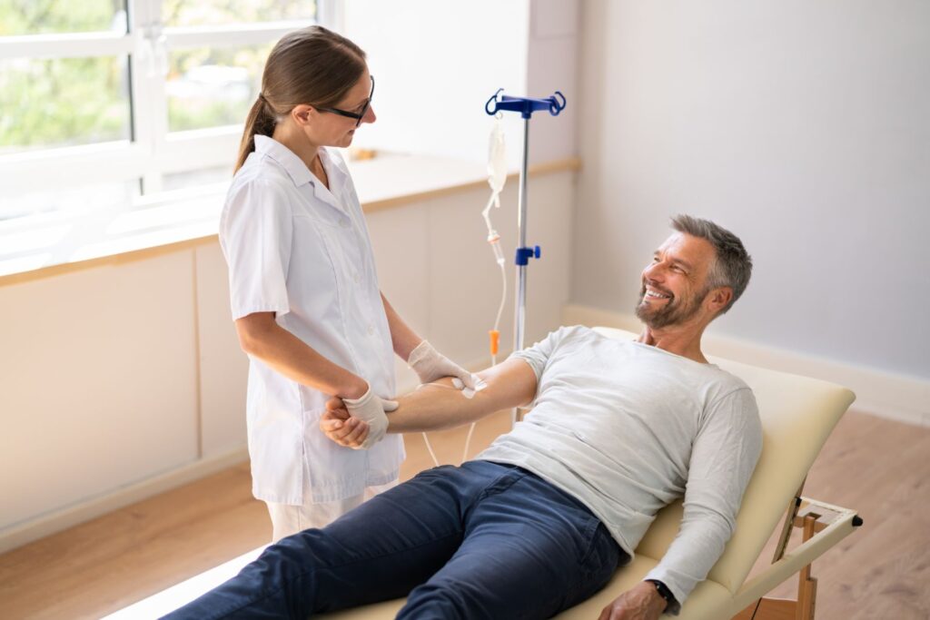 A nurse administers an intravenous (IV) drip to a smiling man lying on a treatment bed in a bright medical room.