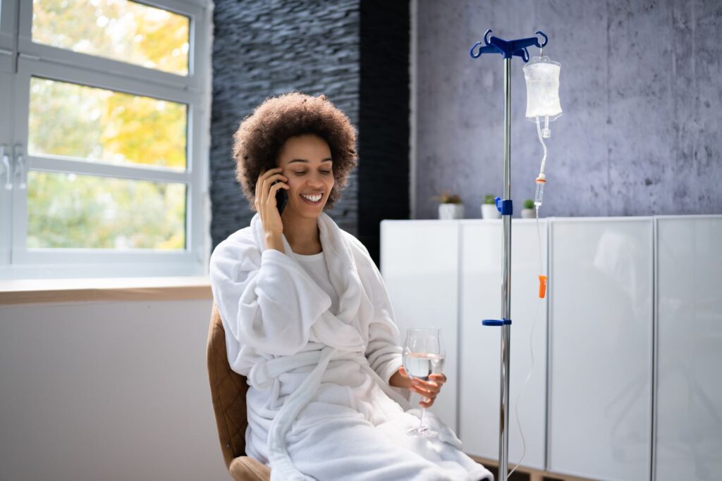 Woman in a white robe sits on a chair, holding a glass of water and talking on the phone, with an IV drip stand beside her in a bright room.