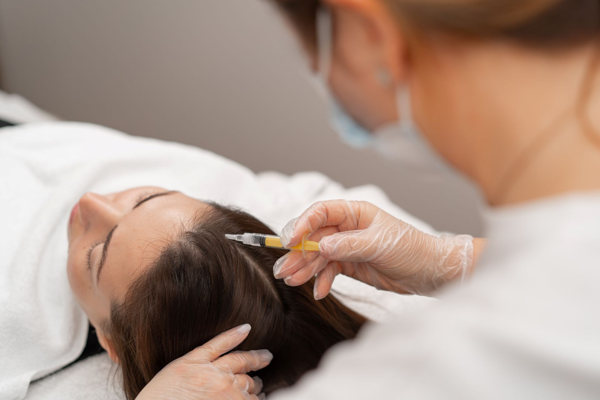 A person receives a Biotin Injection in the scalp while lying on a treatment table. A practitioner wearing gloves and a mask administers the injection.