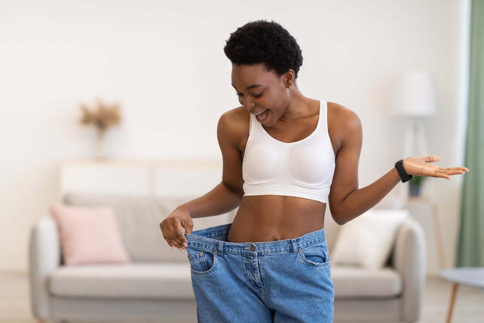 Woman in a white sports bra stands in a living room, smiling and holding out the waistband of oversized jeans to show significant weight loss.