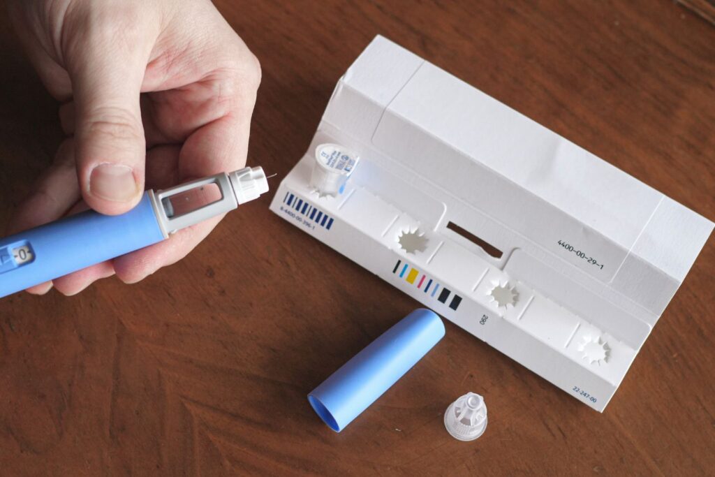 A hand holds an insulin pen beside a white packaging box, pen cap, and two disposable needle tips on a wooden surface.