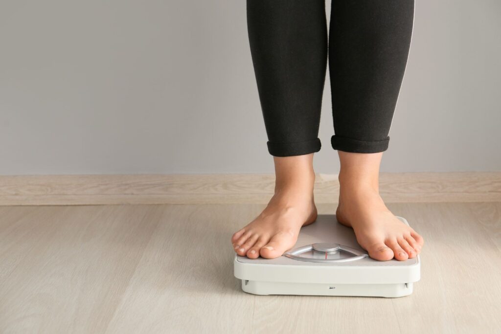 Person wearing black leggings standing barefoot on a white analog bathroom scale on a light wooden floor against a plain wall.