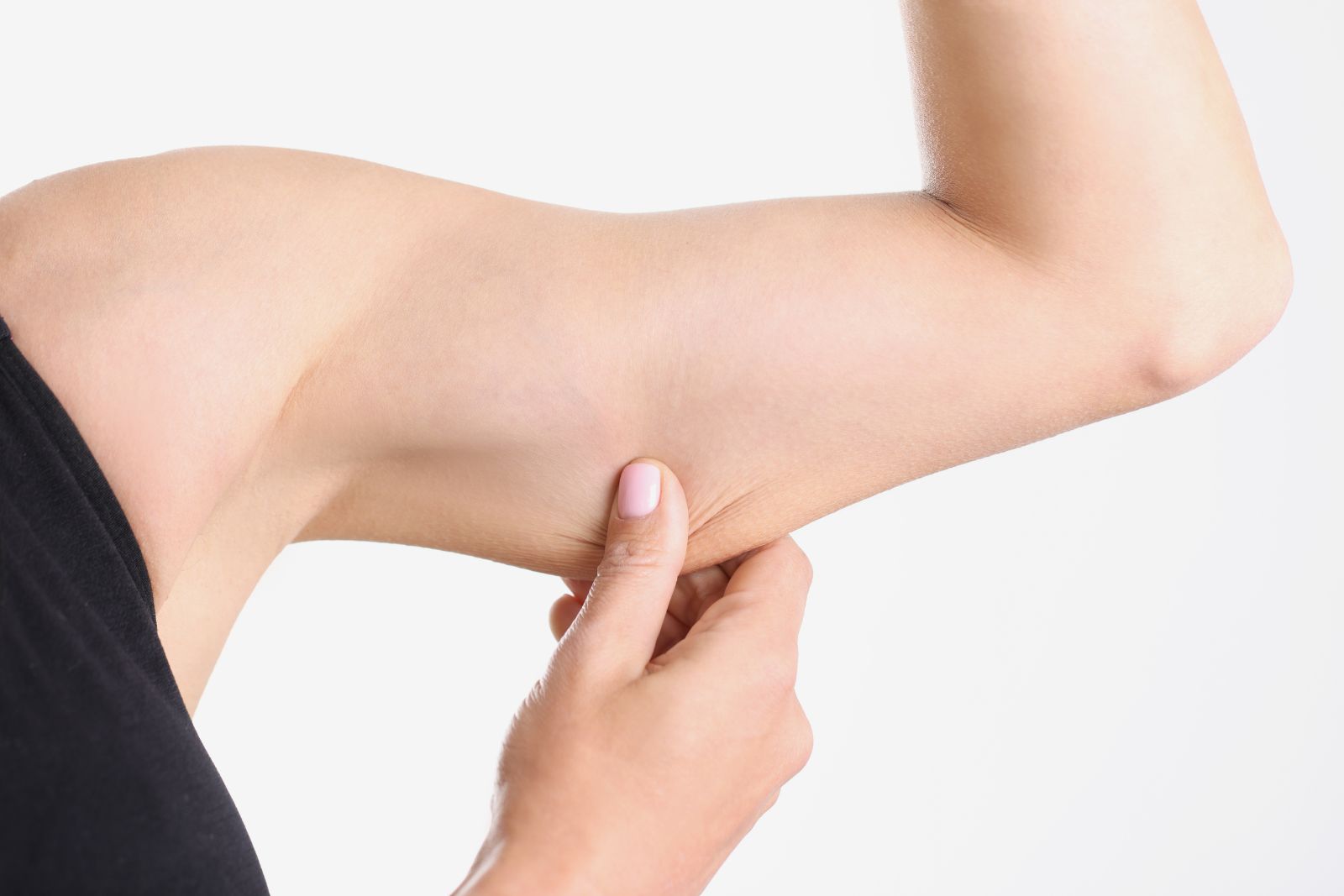 A person pinching the loose skin on the underside of their upper arm, showing a common concern for those wondering how to tighten skin, with their arm bent and wearing a black sleeveless top, against a white background.