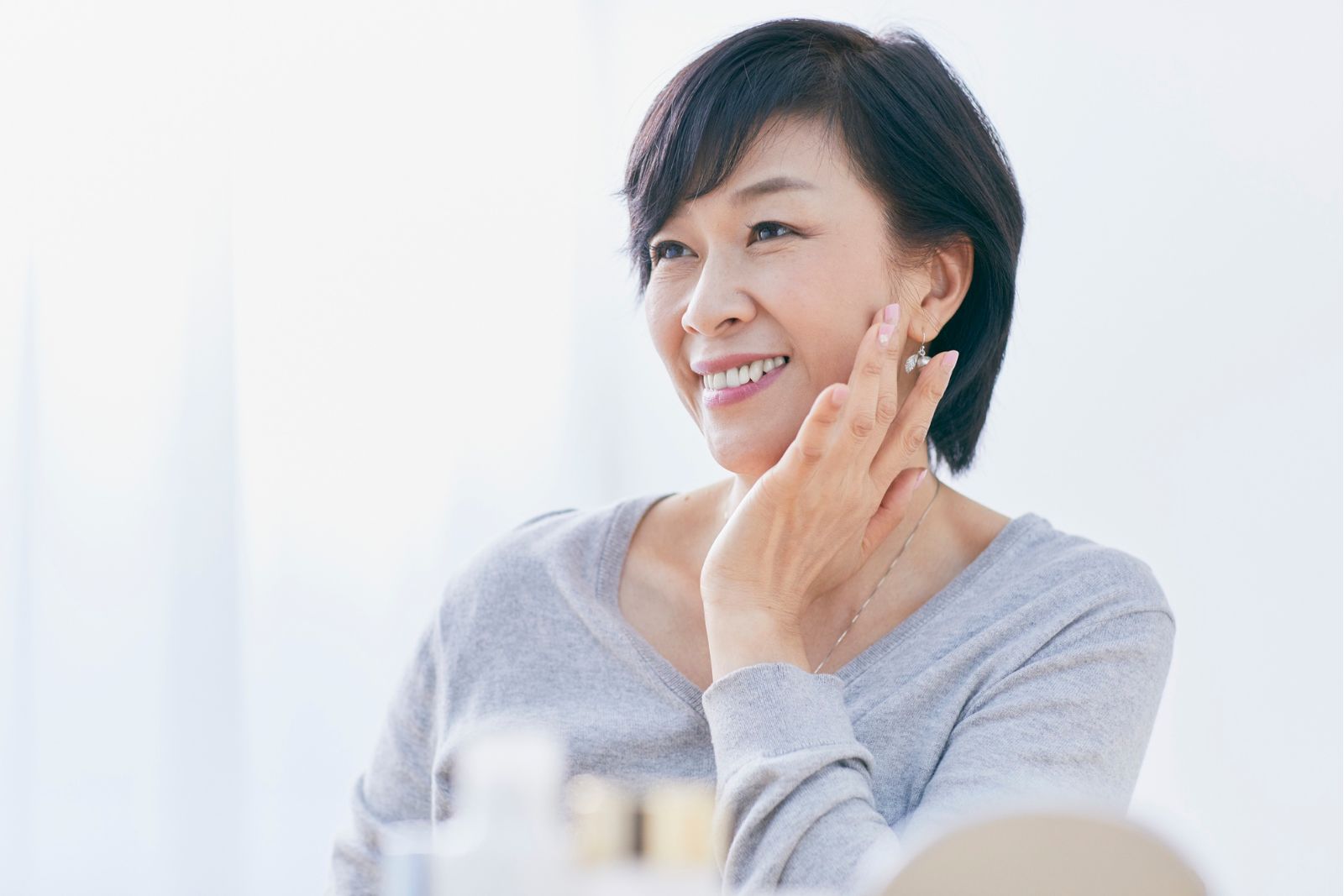 A woman with short dark hair and a light gray top smiles while touching her face, seated indoors with soft lighting—radiating confidence that comes from discovering the best anti-aging treatments.