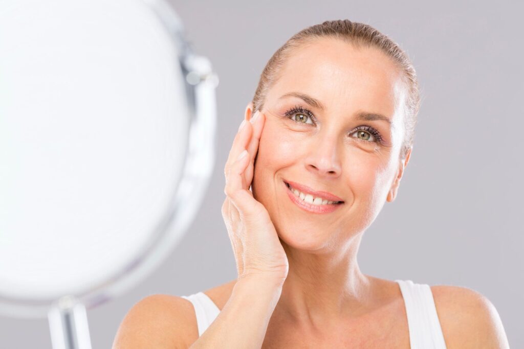 A woman with light brown hair touches her face and smiles while looking at a round mirror in a softly lit room, enjoying one of the best anti aging treatments for 40s.