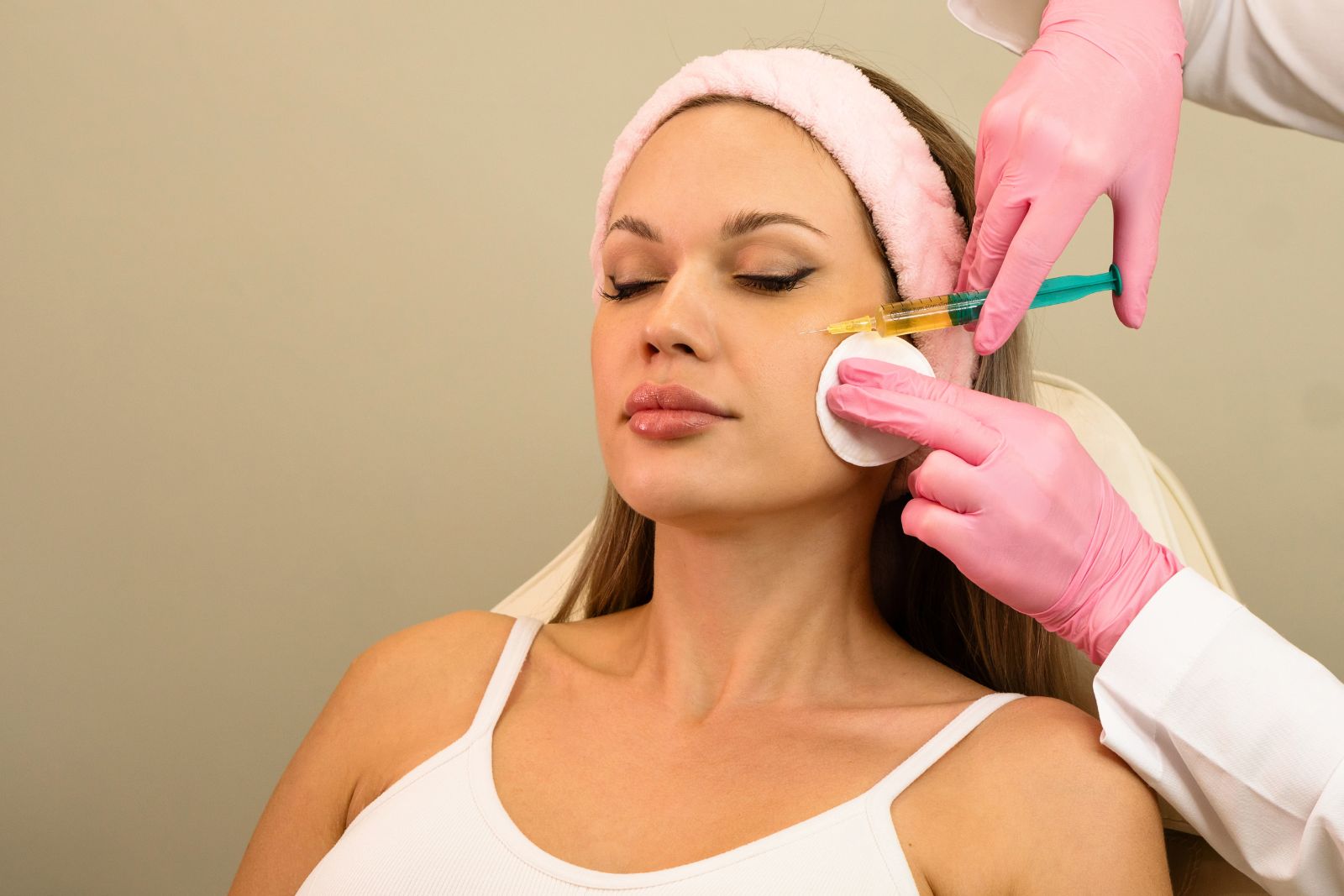 A woman with closed eyes receives a platelet facial injection in her cheek from a gloved professional, while holding a cotton pad to her face.