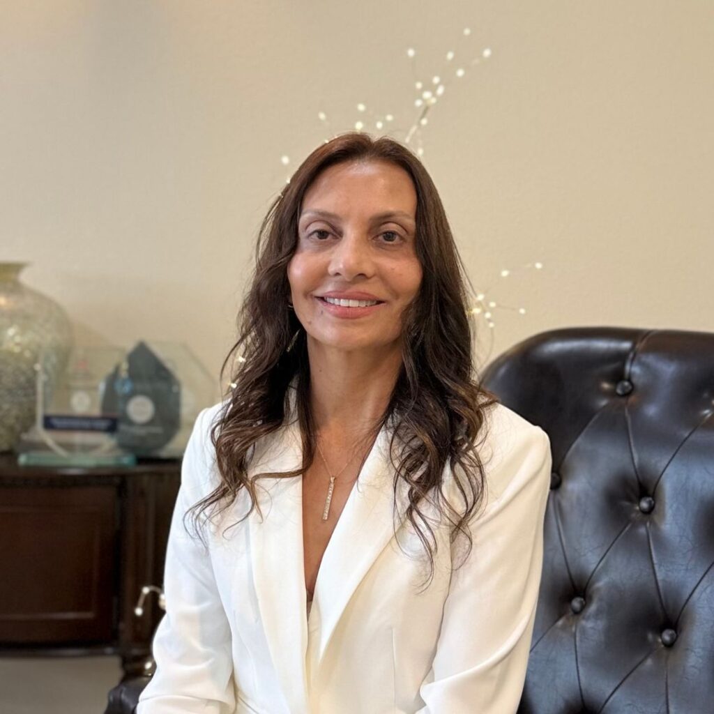 A woman with long brown hair wearing a white blazer sits on a dark leather chair, smiling at the camera, surrounded by the calming neutral decor of Aesthetics Medspa in Reno.