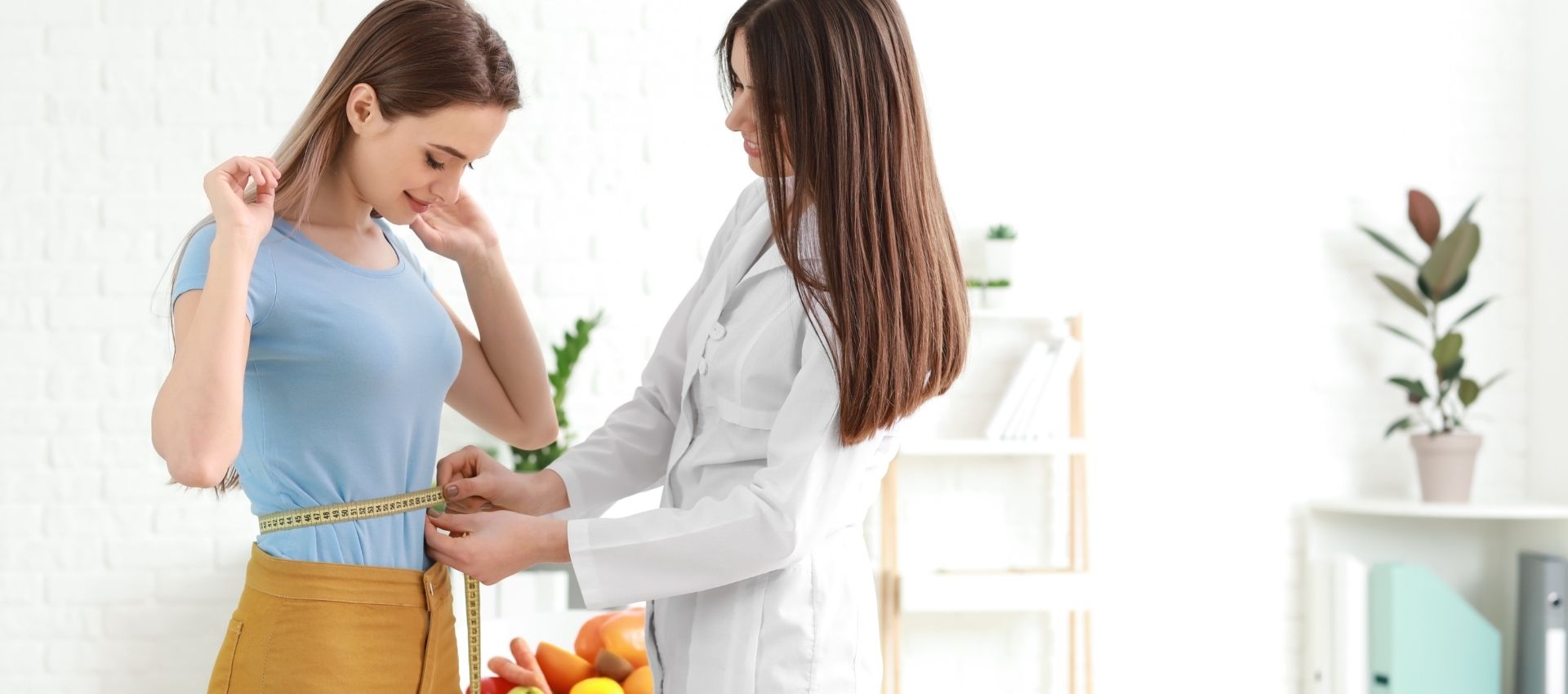A woman in a blue shirt is having her waist measured with a tape measure by a female healthcare professional in a bright office, as part of her zepbound weight loss injections program.