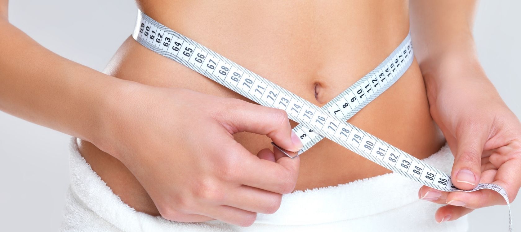 A person measuring their waist with a tape measure, wearing a white towel around their waist, considering Wegovy weight loss injections.