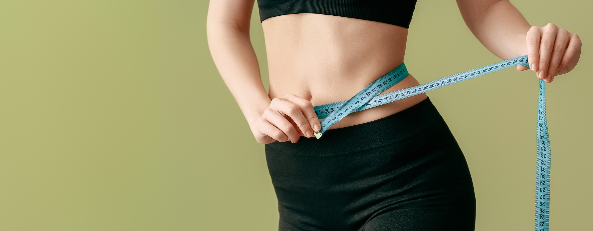Person in black fitness clothing measuring their waist with a blue measuring tape, illustrating progress from a medical weight loss program, against a plain light green background.