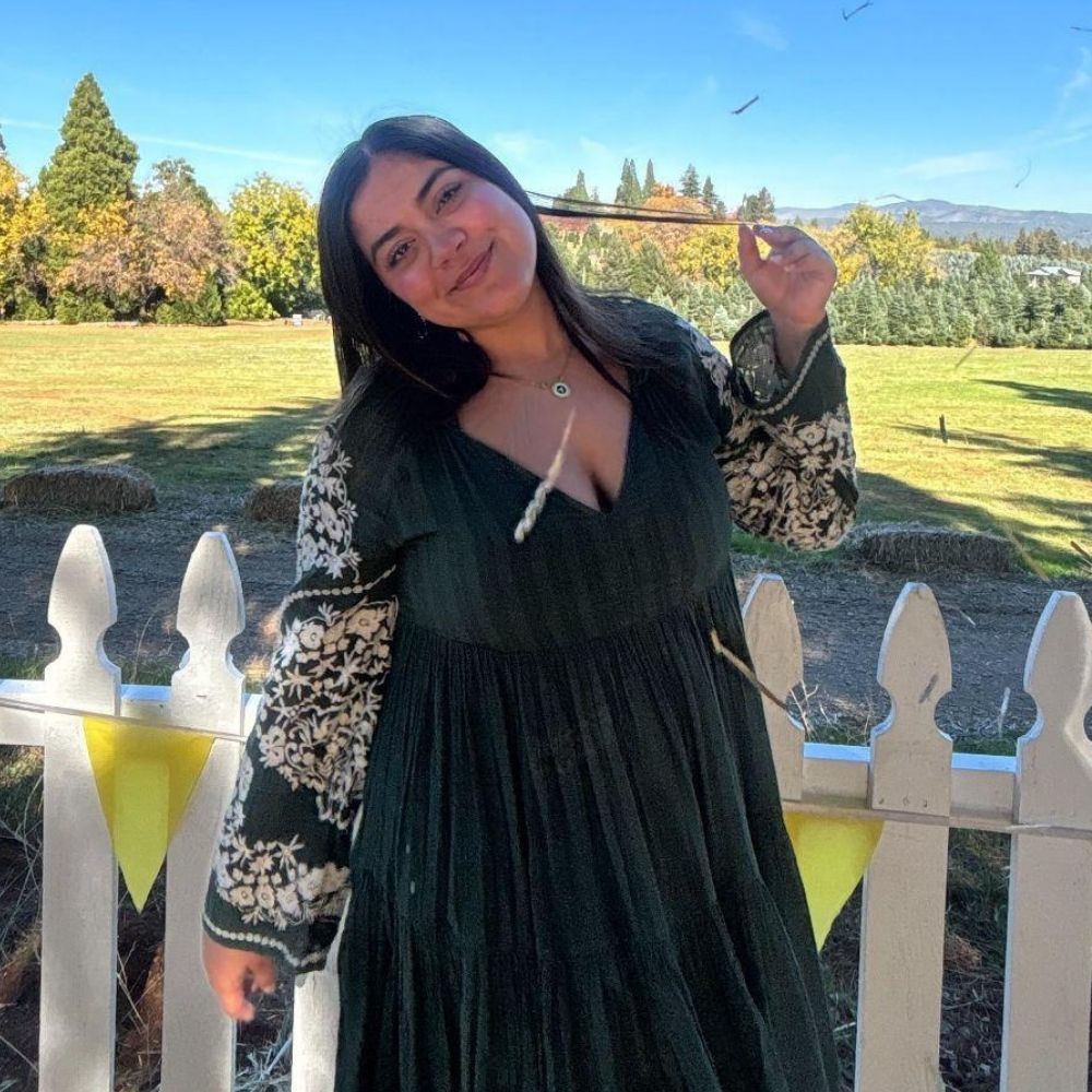 Woman in a dark dress with floral sleeves stands in front of a white picket fence and grassy field, smiling at the camera—radiating confidence like she just stepped out of an Aesthetics Medspa in Reno.