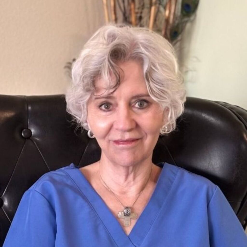 An older woman with short gray hair sits on a black leather chair at a reno Medical Spa, wearing blue scrubs and a necklace, looking at the camera.