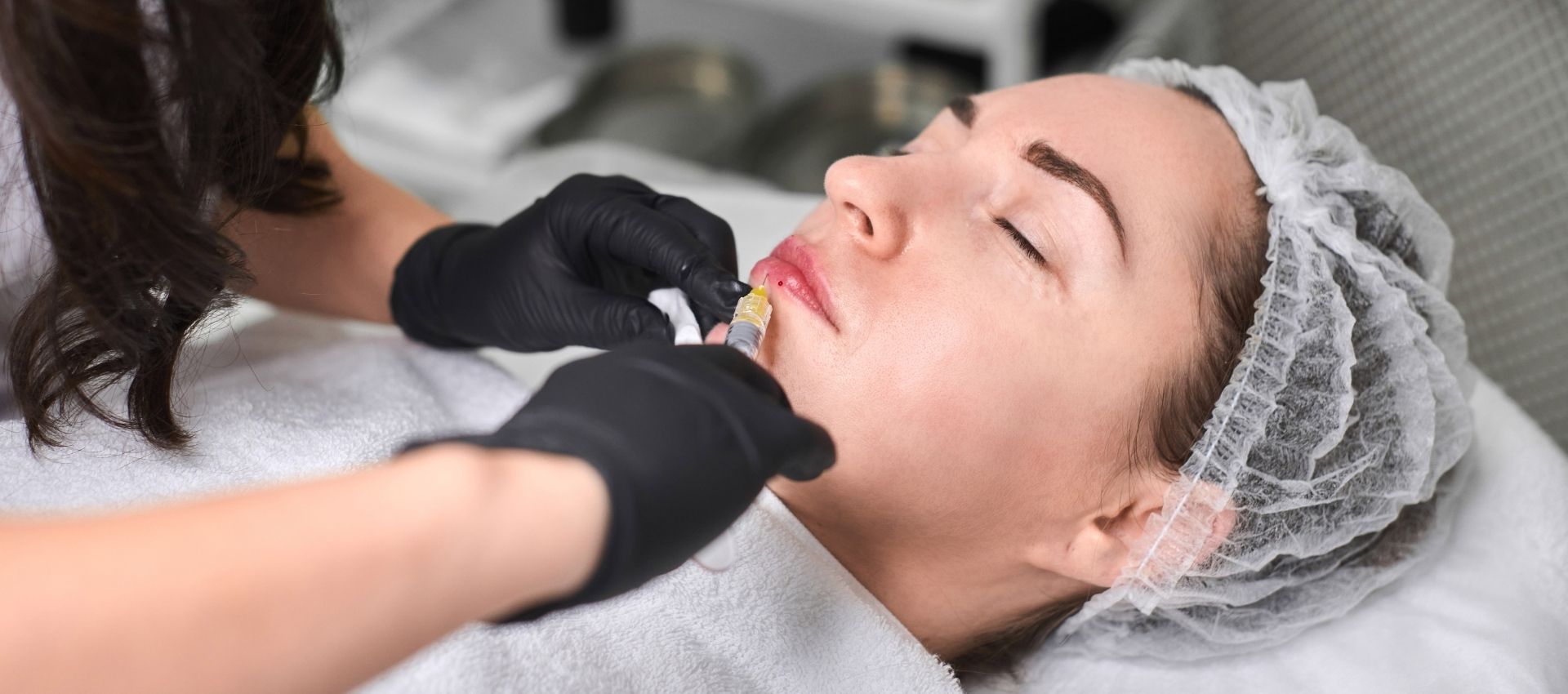 A person with a hair cap lies on a treatment table while a gloved professional performs facial filler injections, enhancing their lips with the benefits of dermal filler injections.