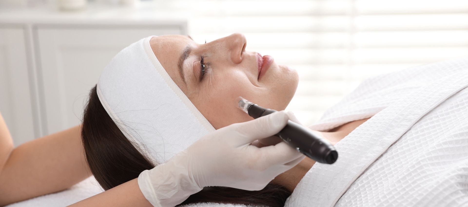 A person receives a PRF Facial treatment with a microneedling device while lying down, wearing a headband and a white robe.