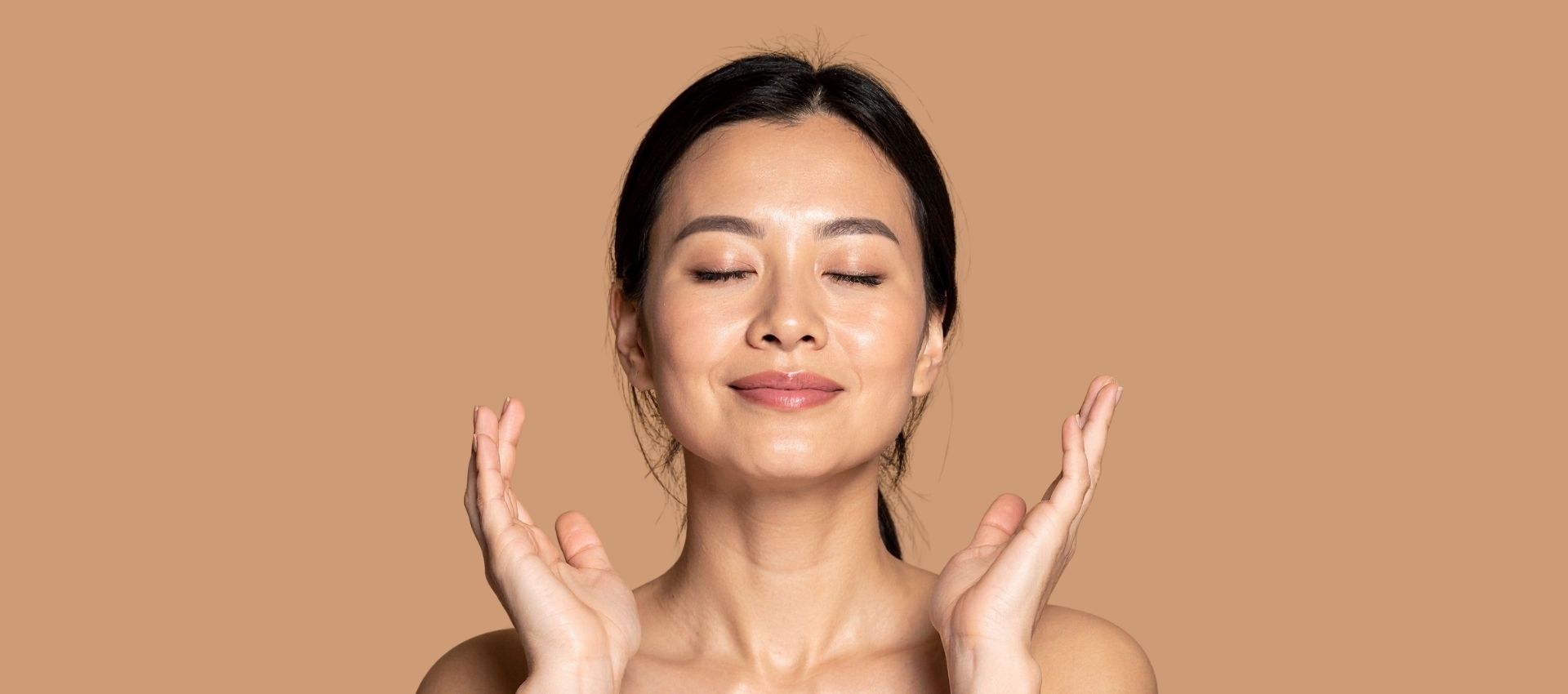 Woman with eyes closed and a slight smile gently touching her face with both hands, posed against a plain beige background—radiant skin suggesting the benefits of PRF Injections.