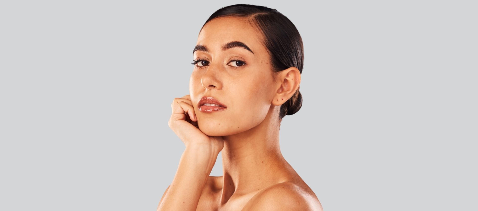A woman with dark hair pulled back looks at the camera with her hand touching her chin, against a plain grey background, subtly highlighting the smooth neckline often achieved with botox for platysmal bands.