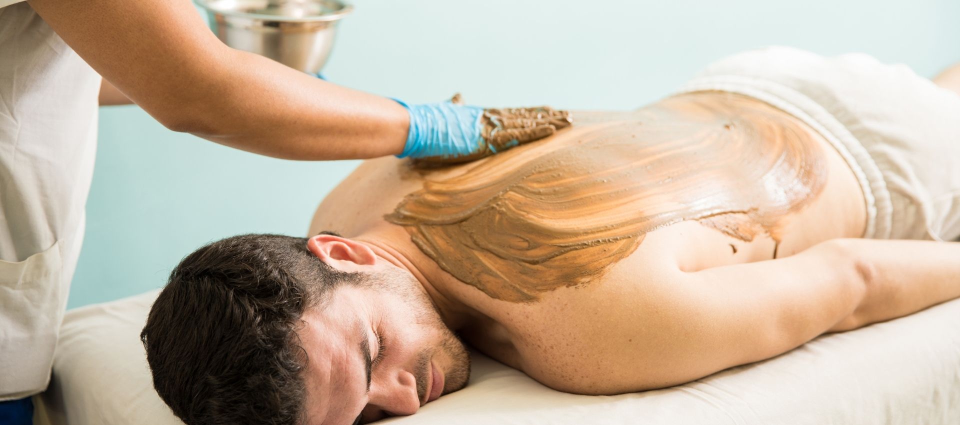 A person lies face down on a massage table while another applies a brown substance to their back with a gloved hand, demonstrating a soothing back facial treatment.