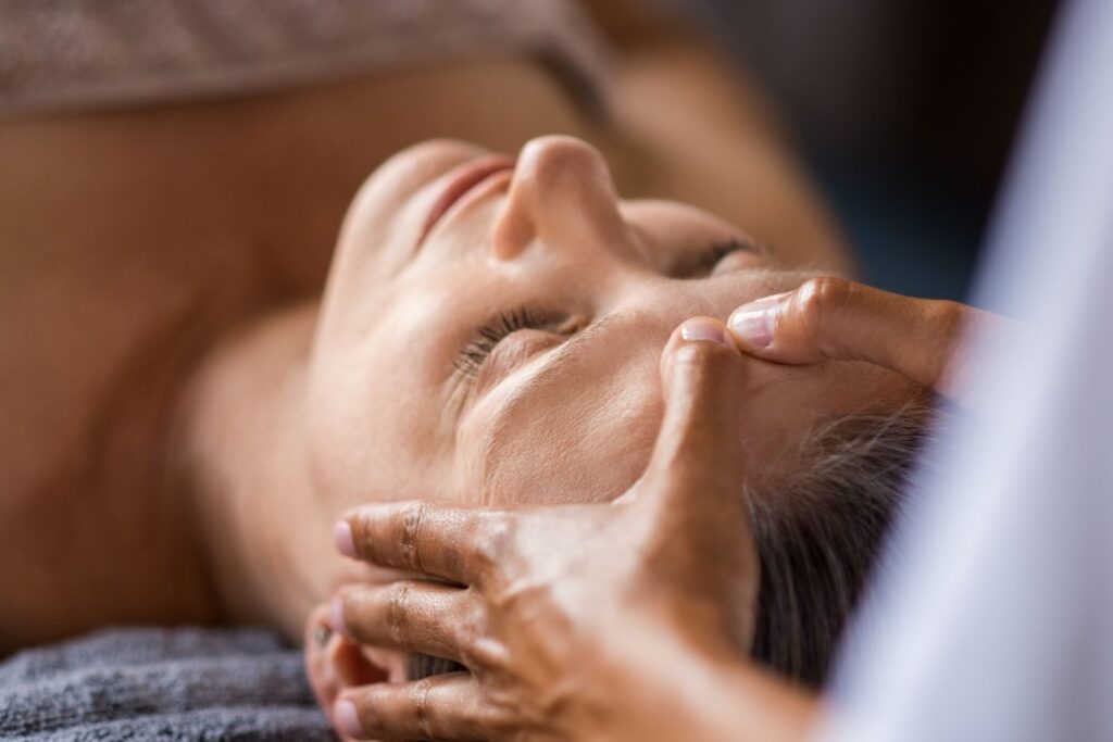 Person lying down with eyes closed receiving a relaxing spa facial treatment, as hands gently press on the temples and forehead area.