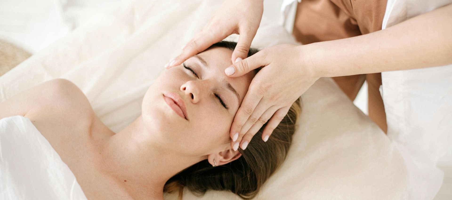 A person lies on a massage table with eyes closed while another gently massages their forehead and temples during a relaxing spa facial treatment.