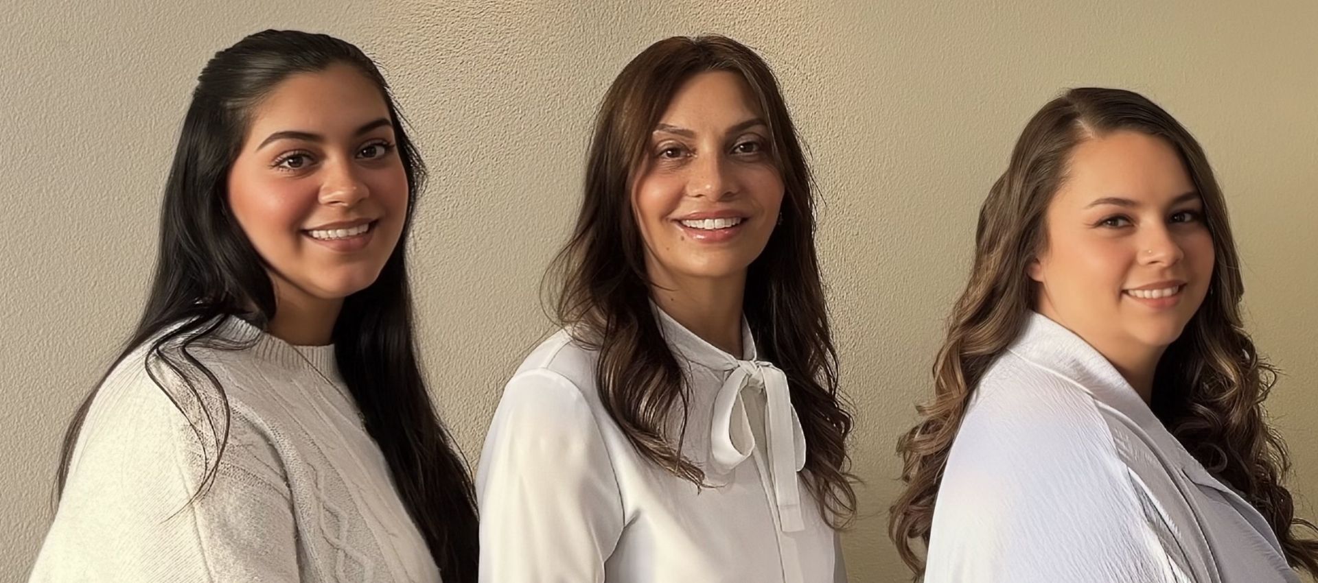 Three women with long hair, wearing white tops, stand side by side in front of a light-colored wall, smiling at the camera—capturing the welcoming atmosphere often found at a top Reno medical spa like Aesthetics Medspa.