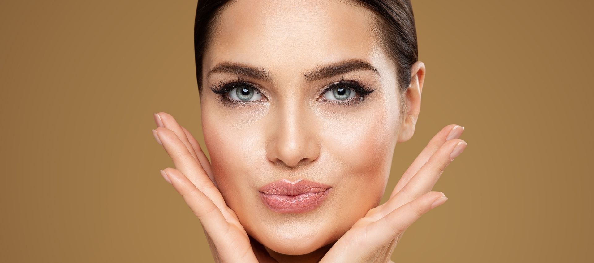 Woman with light eyes and smooth skin poses with her hands framing her face against a beige background, looking directly at the camera with a slight pout, showcasing the subtle elegance that can be achieved through dermal filler injections.