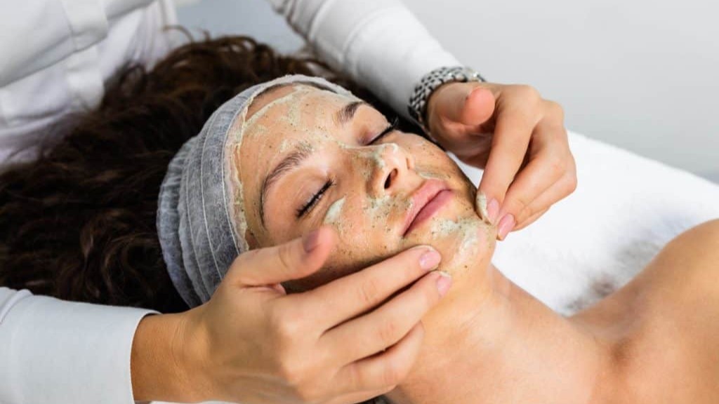 A person receives a relaxing spa facial treatment with a scrub mask applied, while an esthetician gently massages their face.