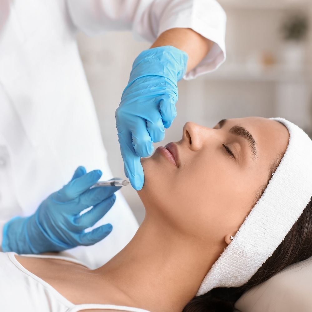 Dermatologist in blue gloves applying a dermal filler injection to a woman's chin.