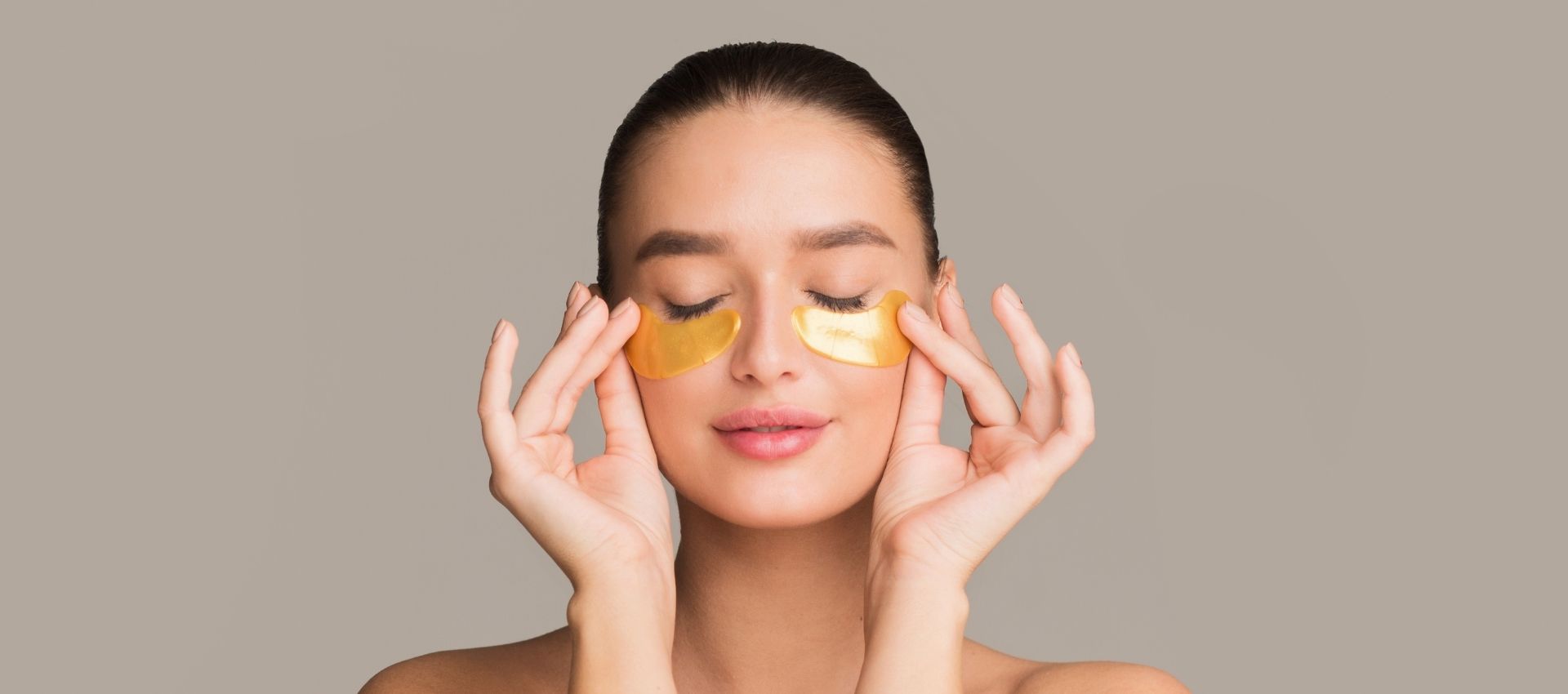 Woman with eyes closed applying gold under-eye patches to her face, mimicking the rejuvenating effects of a tear trough filler, and posing gracefully against a neutral background.